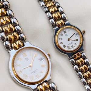 May include: Two Givenchy Paris wristwatches featuring white dials with gold Roman numeral hour markers, gold hands, and a date window. The watches have two-tone link bracelets constructed from alternating silver and gold-toned metal segments.