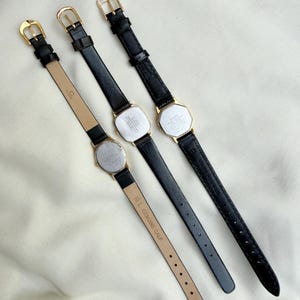 May include: Three vintage gold-toned watches with black leather straps are displayed. The watch faces are rectangular or octagonal. The straps have gold buckles. One strap is stamped "10 L GENUINE CALF". The watches are arranged on a cream-colored surface.