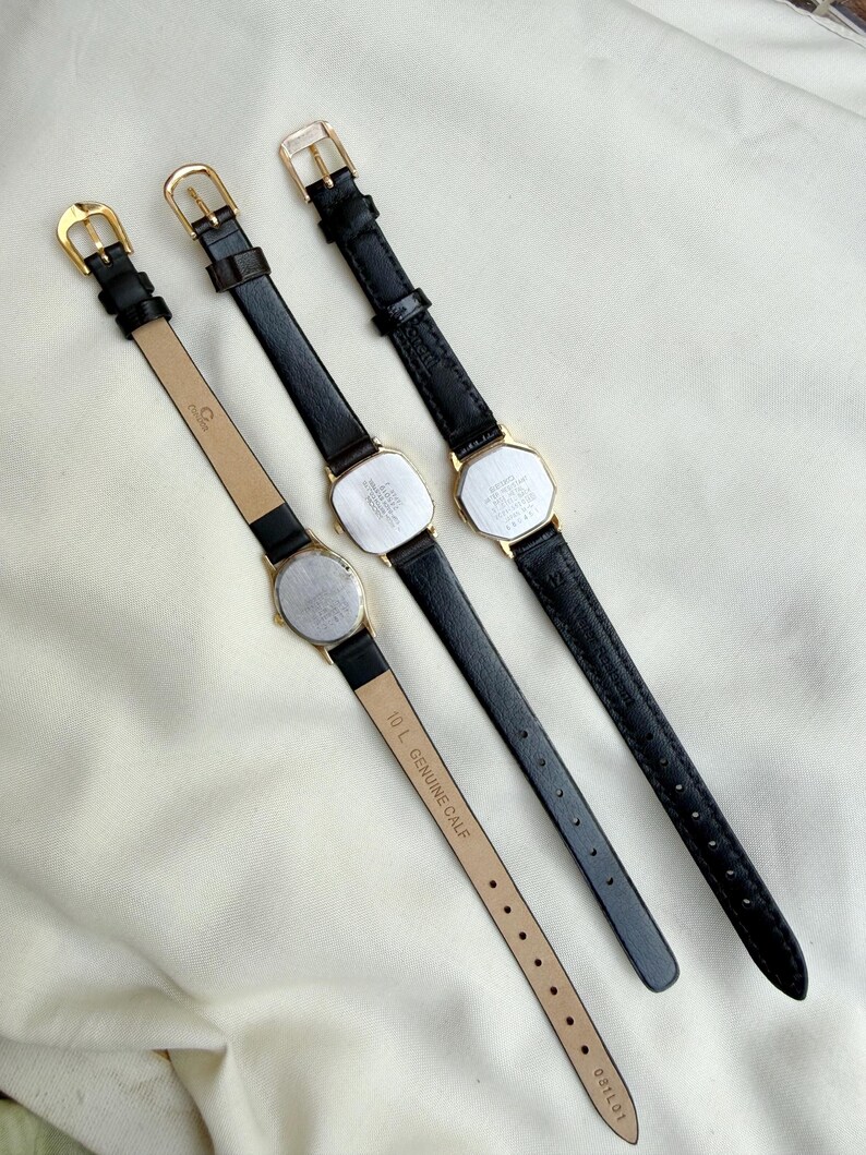 May include: Three vintage gold-toned watches with black leather straps are displayed. The watch faces are rectangular or octagonal. The straps have gold buckles. One strap is stamped "10 L GENUINE CALF". The watches are arranged on a cream-colored surface.