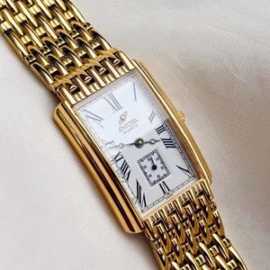 May include: A gold-toned rectangular wristwatch with a white face and black Roman numerals. The watch features a small seconds dial and a gold link band. The watch face displays the brand name "ENICAR QUARTZ".