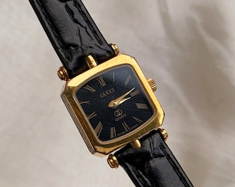 May include: A Gucci quartz wristwatch featuring a square gold-tone case with a black textured dial, gold Roman numeral hour markers, and gold hands. The watch is attached to a black leather strap with a crocodile-embossed pattern and gold lugs.
