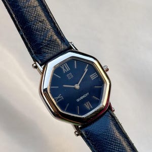 May include: A Givenchy wristwatch with a navy blue face and a silver-colored bezel. The watch has a dark blue leather band. The watch face displays Roman numerals and the Givenchy logo. The watch is an octagonal shape.