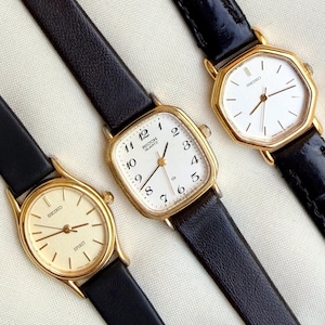 May include: Three vintage gold-toned wristwatches with black leather bands. The watches have white or cream-colored faces with black numerals and hands. One watch face reads "RICOH QUARTZ", and another reads "SEIKO SPIRIT".