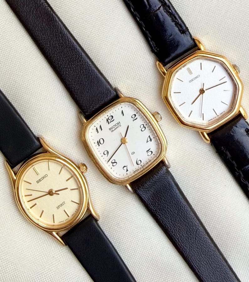 May include: Three vintage gold-toned wristwatches with black leather bands. The watches have white or cream-colored faces with black numerals and hands. One watch face reads "RICOH QUARTZ", and another reads "SEIKO SPIRIT".