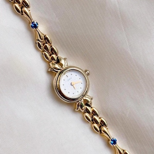 Peut inclure: Une montre-bracelet dorée avec un cadran blanc et des aiguilles dorées. Le bracelet de la montre présente un motif répétitif de maillons dorés et est orné de pierres précieuses bleues. La montre est présentée sur une surface crème.
