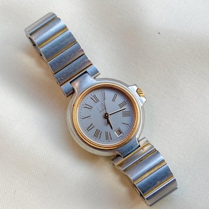 Può includere: Orologio al quarzo Dunhill con cinturino in metallo argentato e dorato. Il quadrante è grigio chiaro con numeri romani e visualizzazione della data. La lunetta e la cassa sono dorate e argentate.