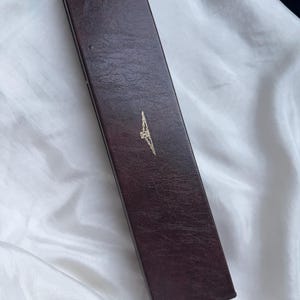 May include: A rectangular, dark brown, textured storage box with a hinged lid. The exterior surface features a faux leather grain finish. Centered on the top lid is a small, gold-colored emblem depicting a winged hourglass logo design.