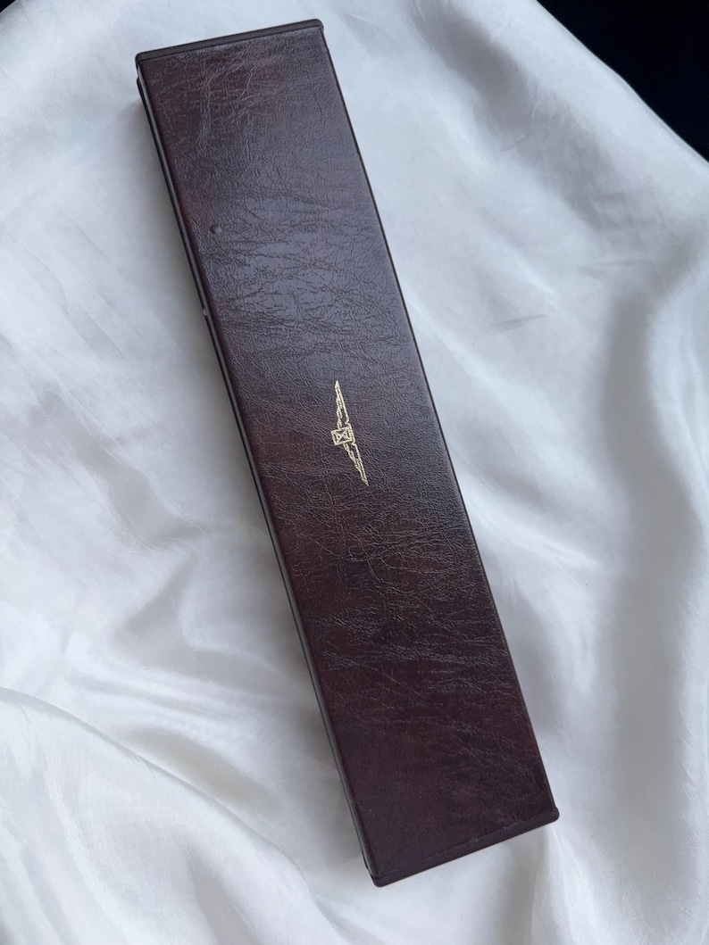 May include: A rectangular, dark brown, textured storage box with a hinged lid. The exterior surface features a faux leather grain finish. Centered on the top lid is a small, gold-colored emblem depicting a winged hourglass logo design.