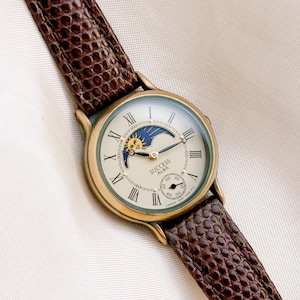 May include: A gold-tone wristwatch with a cream-coloured dial, black Roman numerals, and a brown textured leather strap. The watch face features a sun and moon phase indicator at the top and a small seconds sub-dial at the bottom with the text SUCCESS ALBA.