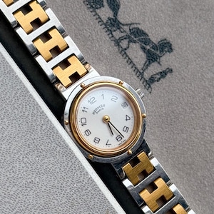 May include: A silver and gold-toned wristwatch with a white face and gold hands. The watch features the brand name "Hermès Paris" and a date display. The watch band has a distinctive H-shaped link design. The watch is presented in a gray box.