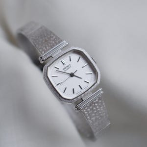 May include: A silver-toned Seiko Quartz watch with a square face and a textured band. The watch has a white dial with black hands and markers. The watch band is made of a woven metal material.