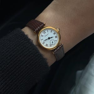 May include: A gold-tone wristwatch with a round white dial featuring black Roman numerals and the Longines brand name. The watch has a textured brown leather strap with a lizard-skin pattern and a small sub-dial located at the six o'clock position.