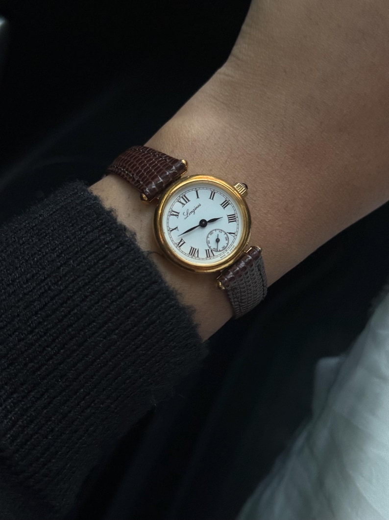 May include: A gold-tone wristwatch with a round white dial featuring black Roman numerals and the Longines brand name. The watch has a textured brown leather strap with a lizard-skin pattern and a small sub-dial located at the six o'clock position.