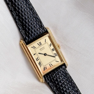 May include: A rectangular gold-toned Seiko Quartz watch with a cream-colored face and black Roman numerals. The watch has a black leather band with a textured pattern. The watch face reads "SEIKO QUARTZ".