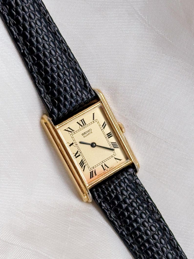 May include: A rectangular gold-toned Seiko Quartz watch with a cream-colored face and black Roman numerals. The watch has a black leather band with a textured pattern. The watch face reads "SEIKO QUARTZ".