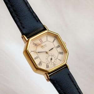May include: A vintage Seiko Quartz watch with a gold-toned octagonal case and a cream-coloured face featuring Roman numeral hour markers. The watch has a small seconds dial and a black leather strap.