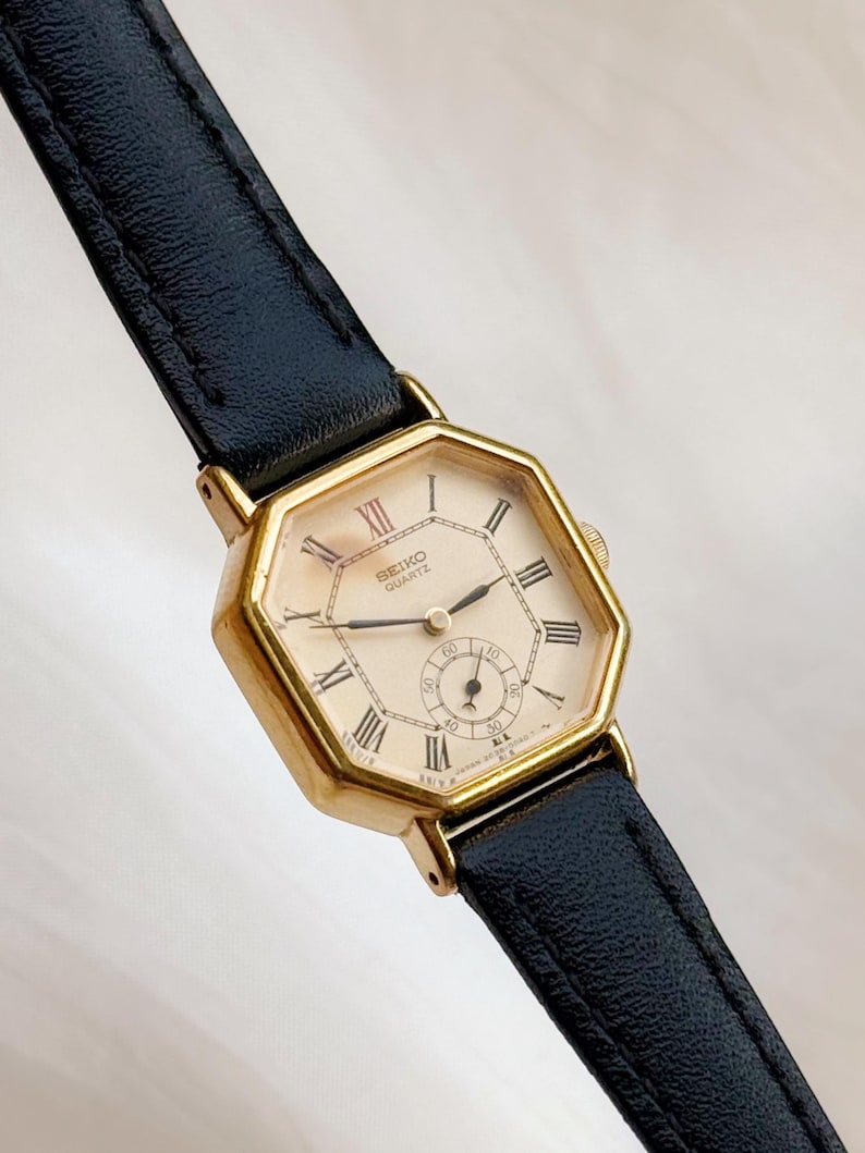 May include: A vintage Seiko Quartz watch with a gold-toned octagonal case and a cream-colored face featuring Roman numeral hour markers. The watch has a small seconds dial and is paired with a black leather strap.
