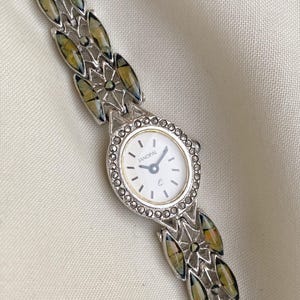 Puede incluir: Un reloj de pulsera Janopal plateado con esfera blanca y bisel decorativo. La correa del reloj presenta detalles ovalados iridiscentes en verde y dorado. Las manecillas indican las 10:09.