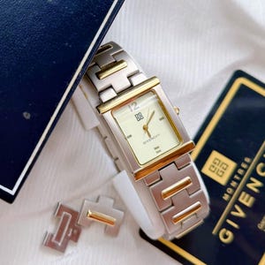 May include: A rectangular Givenchy watch with a silver and gold-tone metal band and case. The watch face is cream-colored with gold hands and hour markers. The watch is displayed with a box and a card.