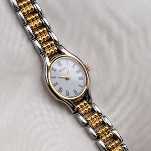 Può includere: Un orologio da polso Seiko con quadrante ovale bianco, numeri romani neri e lancette color oro. L'orologio presenta un cinturino a maglie metalliche bicolore con segmenti alternati argentati e dorati testurizzati e una lunetta color oro.