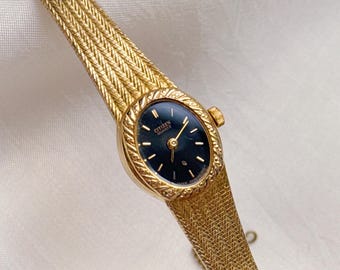 May include: A gold-tone Citizen Quartz watch with an oval face and a dark blue dial. The watch features a textured, woven-style metal band and an ornate, engraved bezel surrounding the dial. The dial displays gold-tone hour markers and hands.