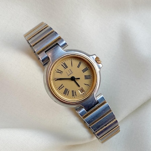 Può includere: Un orologio da polso Dunhill vintage con cinturino in metallo argentato e dorato. Il quadrante dell'orologio è color crema con numeri romani neri e lancette dorate. L'orologio ha un datario a ore 3.