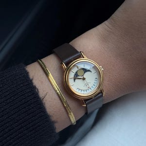 May include: A gold-toned wristwatch with a cream-colored face and a brown leather strap. The watch face features a moon phase design. A gold bracelet is also visible.