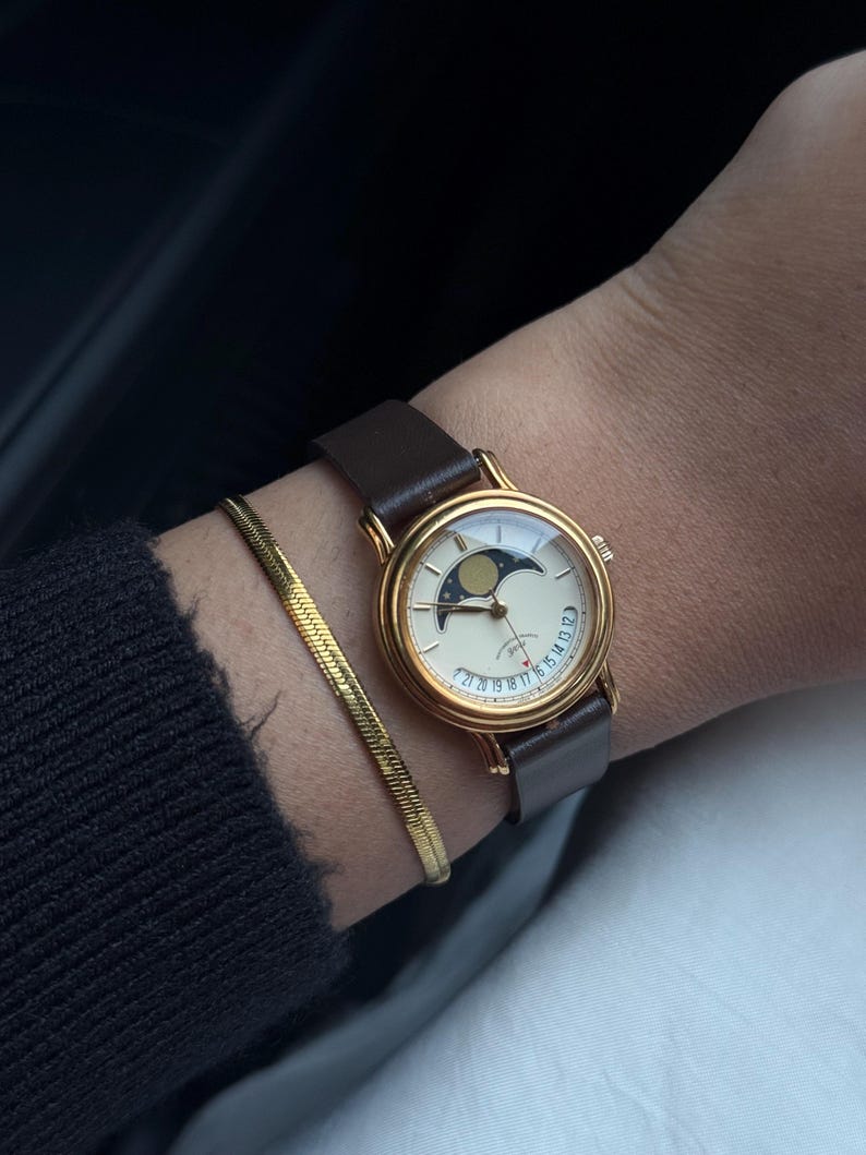 May include: A gold-toned wristwatch with a cream-colored face and a brown leather strap. The watch face features a moon phase design. A gold bracelet is also visible.