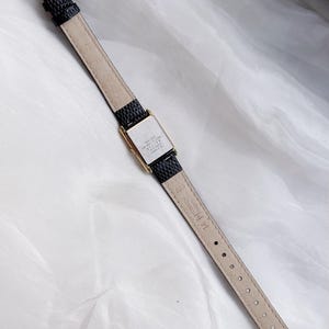 May include: A rectangular gold-toned watch with a black textured leather band. The watch face is silver with black text. The band has a gold-toned buckle. The watch is displayed on a white, textured surface.