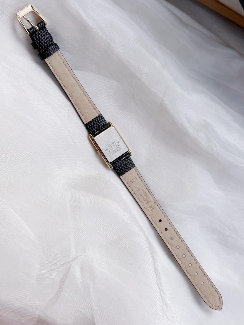 May include: A rectangular gold-toned watch with a black textured leather band. The watch face is silver with black text. The band has a gold-toned buckle. The watch is displayed on a white, textured surface.