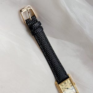 May include: A gold-toned rectangular wristwatch with a black textured leather band. The watch face has Roman numerals and gold hands. The buckle is gold and has the brand name Seiko.