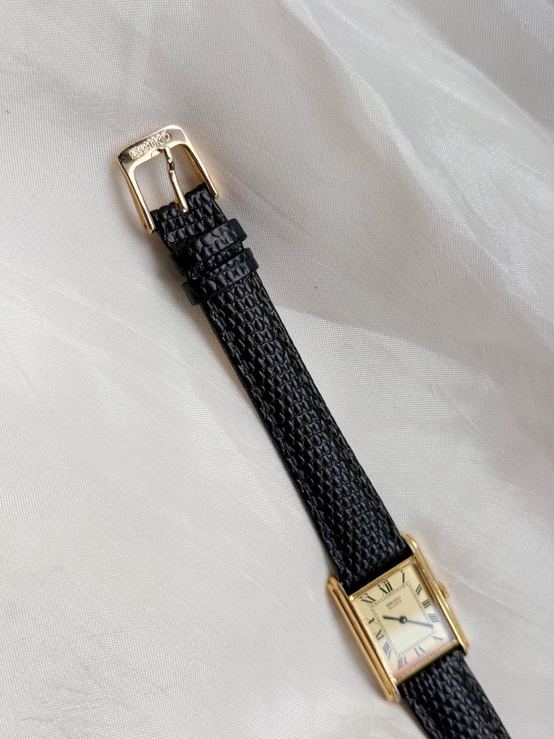 May include: A gold-toned rectangular wristwatch with a black textured leather band. The watch face has Roman numerals and gold hands. The buckle is gold and has the brand name Seiko.