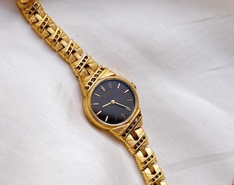 Puede incluir: Un reloj de pulsera dorado con una esfera redonda negra que presenta marcadores de hora y manecillas doradas simples. El reloj tiene una correa de eslabones metálicos texturizados con un patrón entrecruzado que sale de la caja.