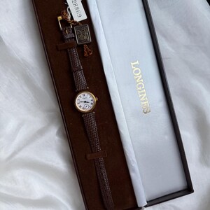 May include: A gold-tone Longines wristwatch with a round white dial, black Roman numerals, and a textured dark brown leather strap. The watch is presented in a rectangular brown and white box with the Longines logo and a small tag labeled G 28869.