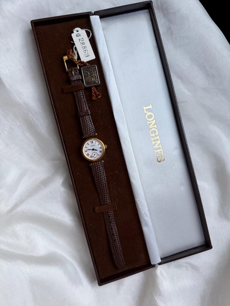 May include: A gold-tone Longines wristwatch with a round white dial, black Roman numerals, and a textured dark brown leather strap. The watch is presented in a rectangular brown and white box with the Longines logo and a small tag labeled G 28869.