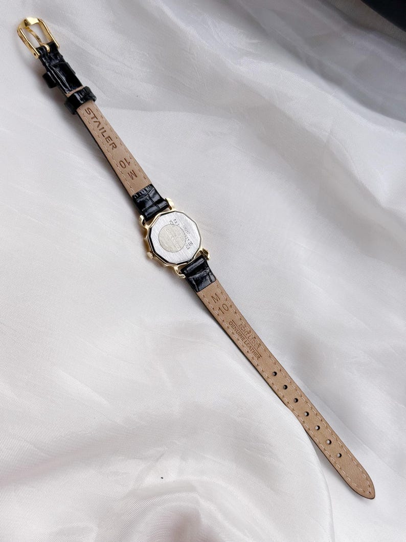 May include: A gold wristwatch with a black leather strap. The watch face is octagonal. The strap is marked "STAIER" and "GENUINE LEATHER". The watch is shown on a white, textured background.
