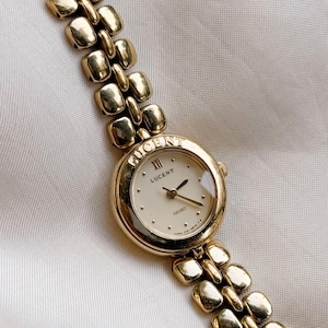Könnte beinhalten: Eine goldfarbene Lucent Seiko Armbanduhr mit cremefarbenem Zifferblatt und goldenen Zeigern. Die Uhr hat ein rechteckiges Gliederarmband. Das Zifferblatt zeigt das Wort "LUCENT" und römische Ziffern. Die Uhr liegt auf einer weißen Satin-Oberfläche.