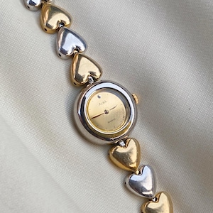 May include: A vintage Alba quartz watch with a gold-toned face and silver-toned hands. The watch has a silver and gold-toned heart-shaped link band. The watch face is round with the brand name "Alba" printed on it.