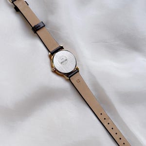 May include: A gold-toned wristwatch with a black leather band and a beige underside. The watch face is round with a silver-colored back. The band is fastened with a gold-colored buckle. The watch is lying on a white, textured surface.