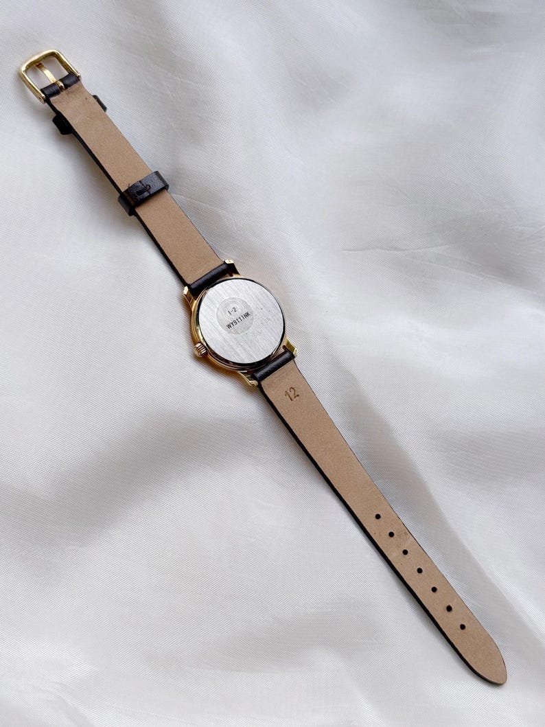 May include: A gold-toned wristwatch with a black leather band and a beige underside. The watch face is round with a silver-colored back. The band is fastened with a gold-colored buckle. The watch is lying on a white, textured surface.