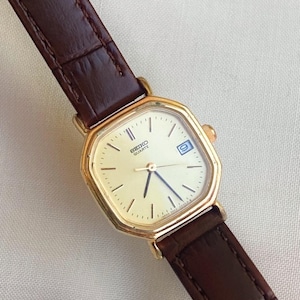 May include: A gold Seiko Quartz watch with a rectangular face and a brown leather strap. The cream-coloured watch face has black hour markers and blue hands. The watch also features a date display at the 3 o'clock position.