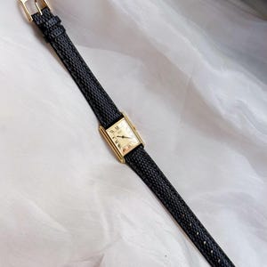 May include: A rectangular gold-toned wristwatch with a cream-colored face and black leather band. The watch features Roman numeral hour markers and a gold buckle. The band has a textured pattern.