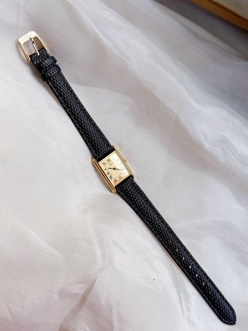 May include: A rectangular gold-toned wristwatch with a cream-colored face and black leather band. The watch features Roman numeral hour markers and a gold buckle. The band has a textured pattern.