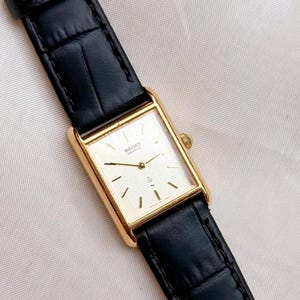 Può includere: Un orologio da polso rettangolare Seiko Quartz color oro con quadrante bianco, indici e lancette sottili color oro. L'orologio è dotato di un cinturino in pelle nera con motivo a trama di coccodrillo e una fibbia color oro.