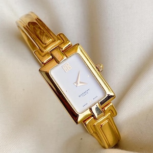 Puede incluir: Un reloj Givenchy Paris dorado con una esfera blanca rectangular. El reloj tiene una correa dorada y una pequeña corona. La esfera muestra el logotipo de Givenchy y las palabras "SWISS MADE".
