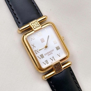 May include: A rectangular gold-tone wristwatch with a white face, black Roman numerals, and gold hands. The dial features the Givenchy Paris logo and a small square emblem. It has a black leather strap with gold-tone lugs featuring the Givenchy logo.