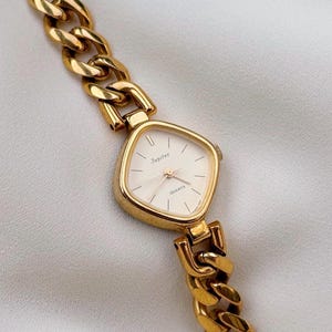 May include: A gold-tone analog wristwatch featuring a cushion-shaped case and a thick curb chain link bracelet. The white dial displays the text Jupiter and QUARTZ, with thin gold-tone hour markers and matching hands for a clean, classic appearance.