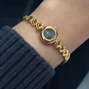 May include: A gold-tone Seiko wristwatch featuring a small, round black dial with a single diamond marker at the twelve o'clock position. The watch has a polished gold-tone metal link bracelet with a repeating interlocking pattern design.
