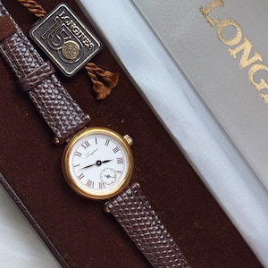 May include: A gold-tone Longines wristwatch with a white dial, black Roman numerals, and a small sub-dial. The watch features a textured brown leather strap and is accompanied by a square tag marked Longines 150 1832-1982, presented in a box.