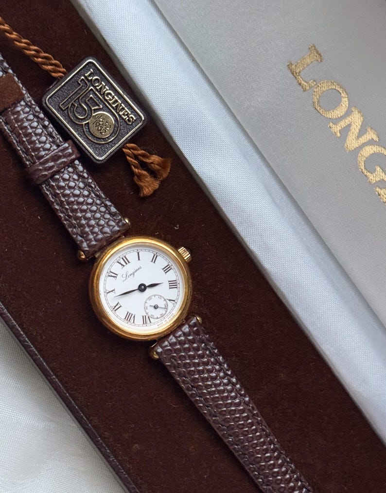 May include: A gold-tone Longines wristwatch with a white dial, black Roman numerals, and a small sub-dial. The watch features a textured brown leather strap and is accompanied by a square tag marked Longines 150 1832-1982, presented in a box.